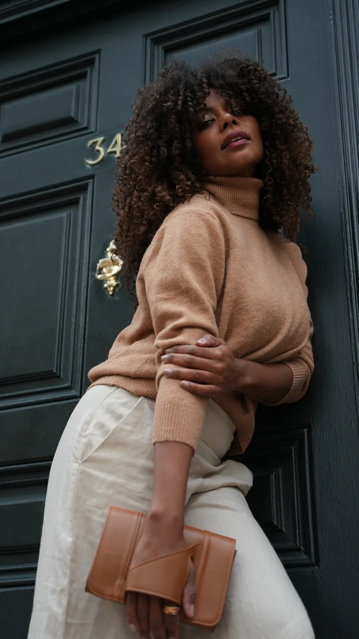 Mujer con sweater camel y clutch de cuero café junto a puerta oscura